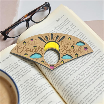 Bookmark with 'Head in the clouds, nose in a book' text on an open book with glasses and a cup.