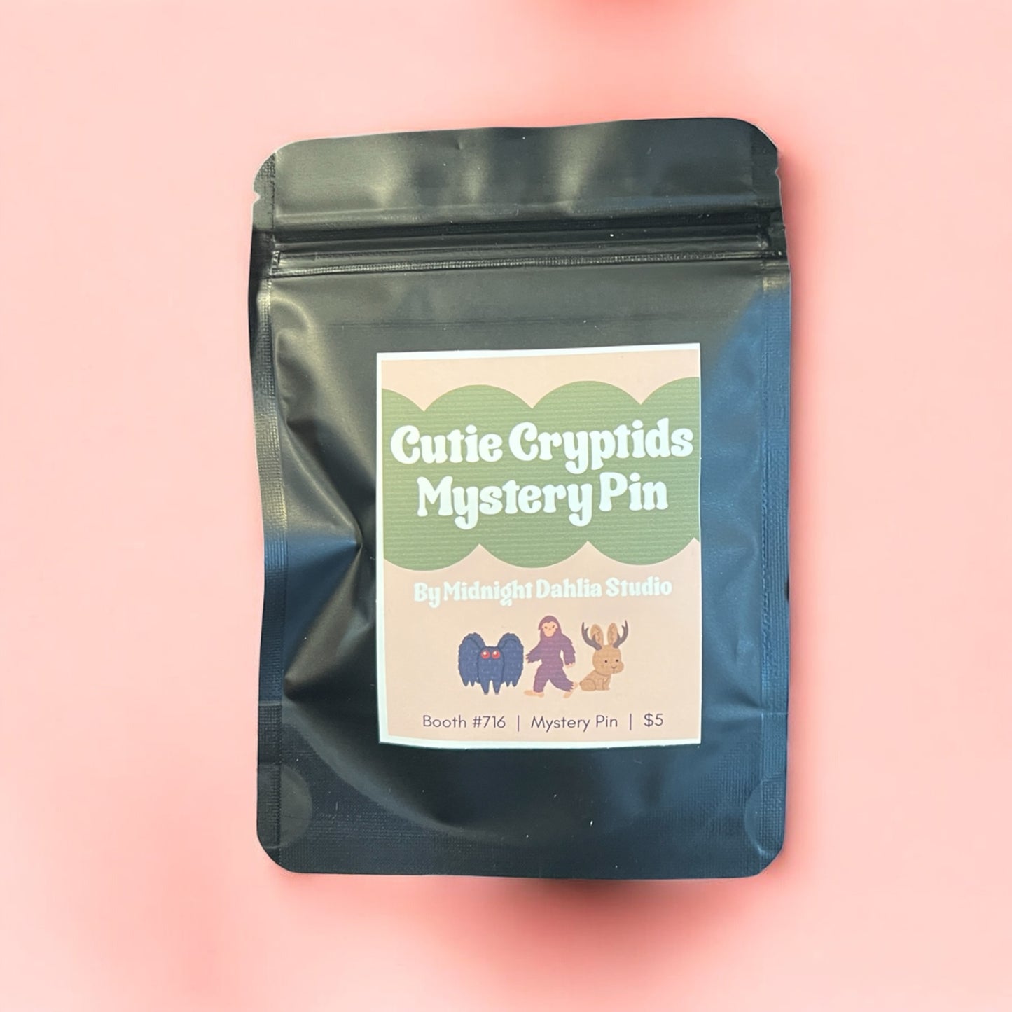 Black ziplock bag with 'Cutie Cryptids Mystery Pin' label on a pink background