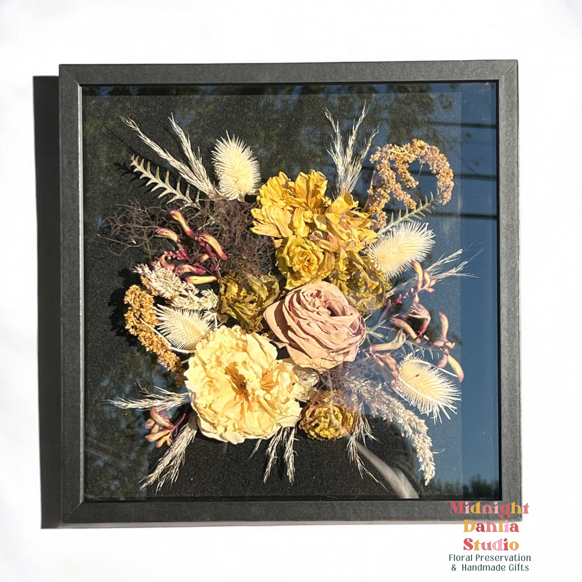 a wooden framed shadow box filled with many flowers, including white roses, pink roses, yellow dahlias, pink red kangaroo paw, pampas grass, white bleached thistle, and leaves.