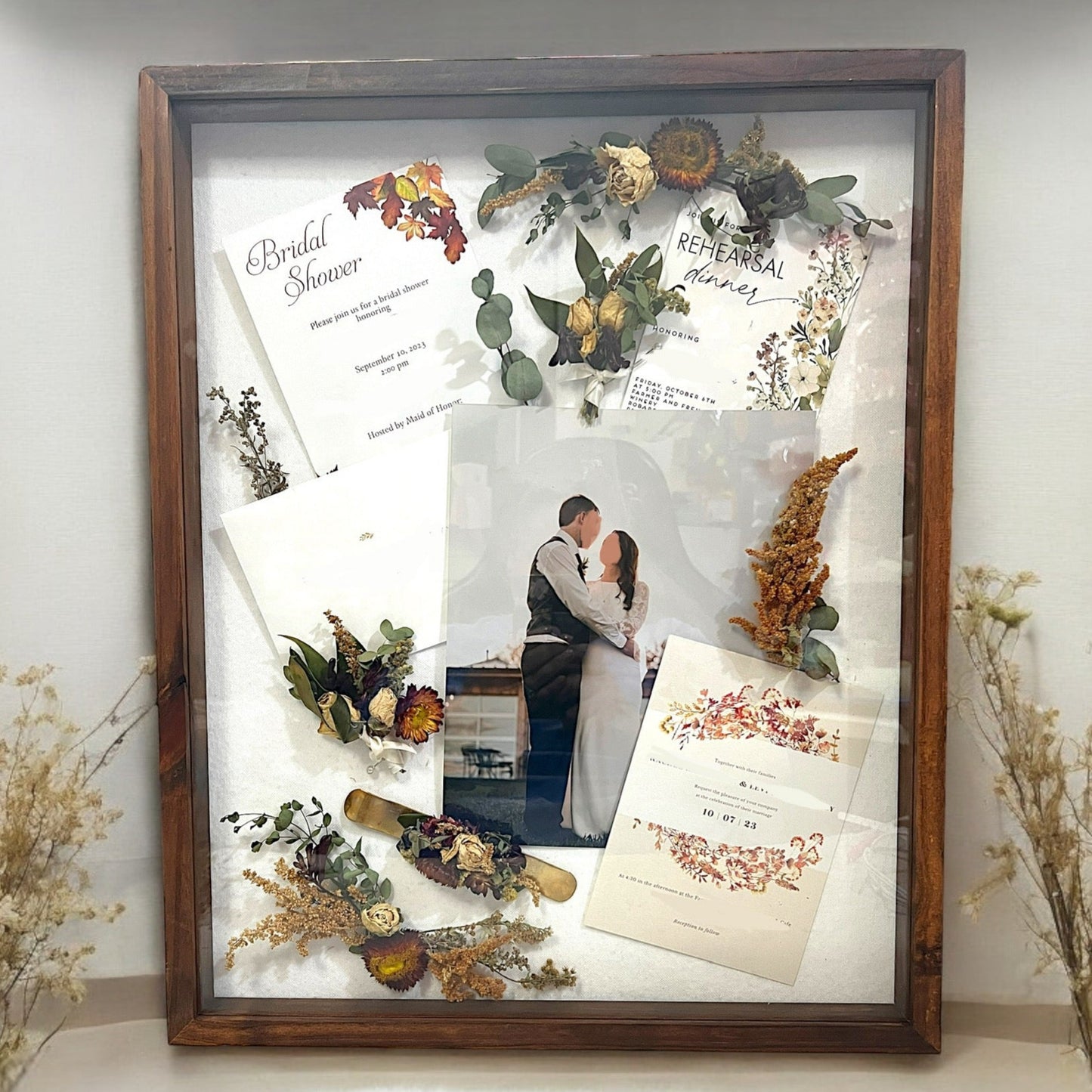 a wooden framed shadow box filled with many small flowers with a mother's corsage, a father's boutonniere and mementos, including a wedding invitation, announcement, photograph of bride and groom, bridal shower invitation, and wedding program.