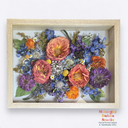 a wooden framed shadow box filled with many flowers, including peach pink roses, purple zinnia, blue delphinium, orange ranunculus, purple carnations, and green leaves.