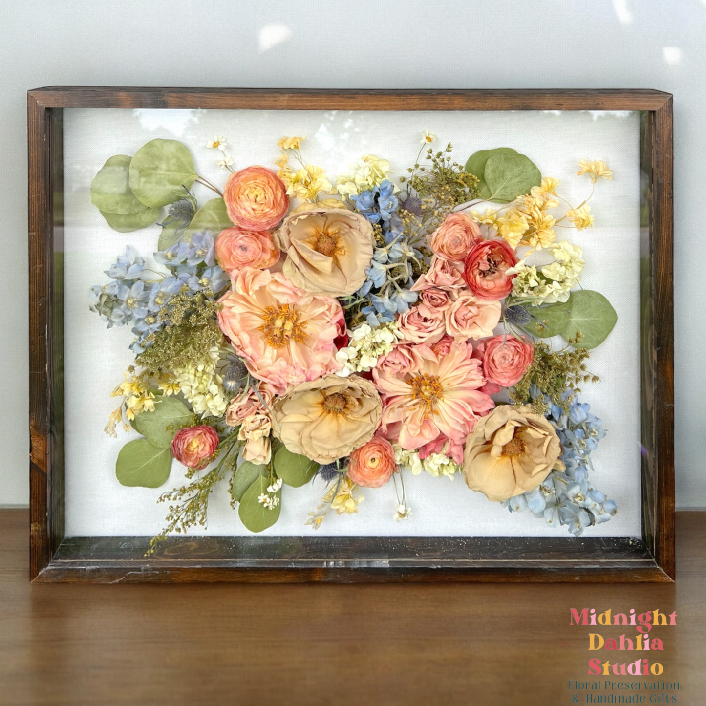 a wooden framed shadow box filled with many flowers, including peach peonies, taupe roses, white stock, blue delphinium, coral peach ranunculus, white stock, white hydrangea, yellow solidago, and silver dollar eucalyptus