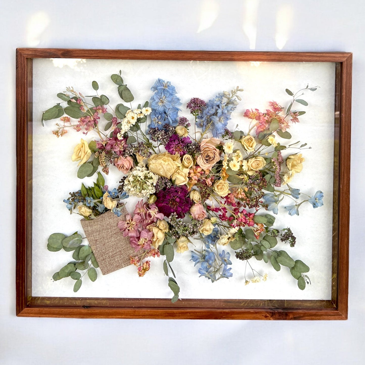 a wooden framed shadow box filled with many flowers, including peach mini spray roses, pink stock, yellow and white roses, blue delphinium, and green leaves.