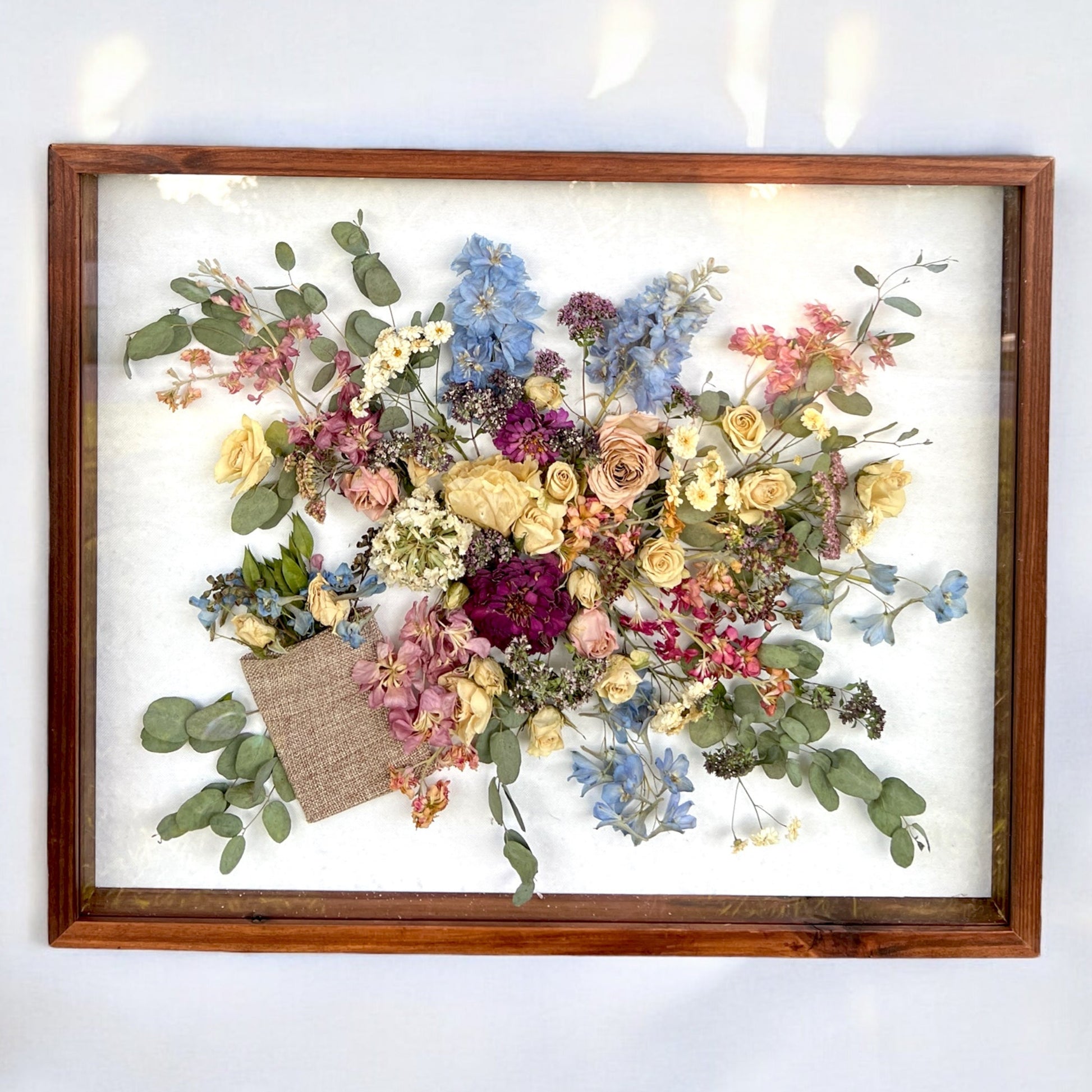 a wooden framed shadow box filled with many flowers, including peach mini spray roses, pink stock, yellow and white roses, blue delphinium, and green leaves.