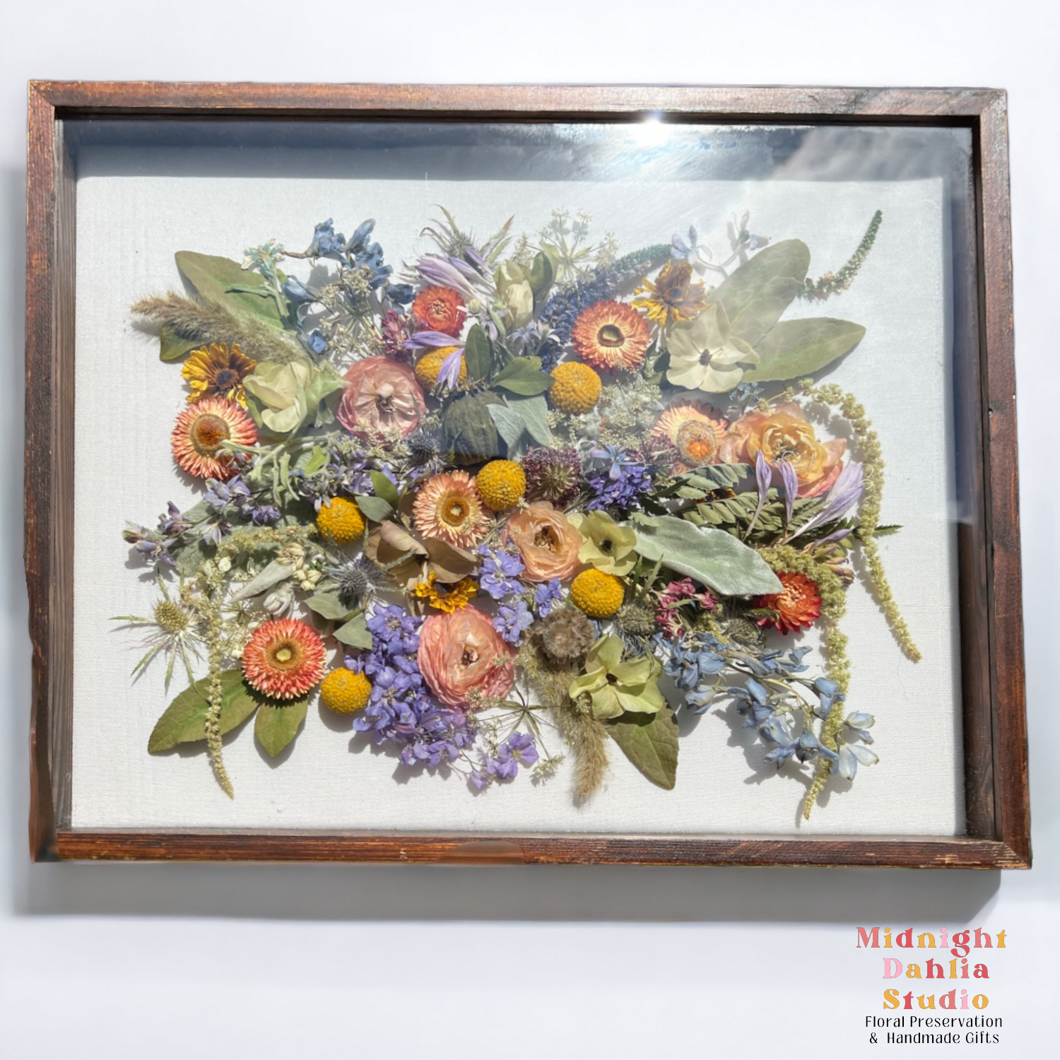a wooden framed shadow box filled with many flowers, including peach ranunculus, strawflowers, blue delphinium, green amaranthus, yellow billy balls, blue thistle, and green leaves.