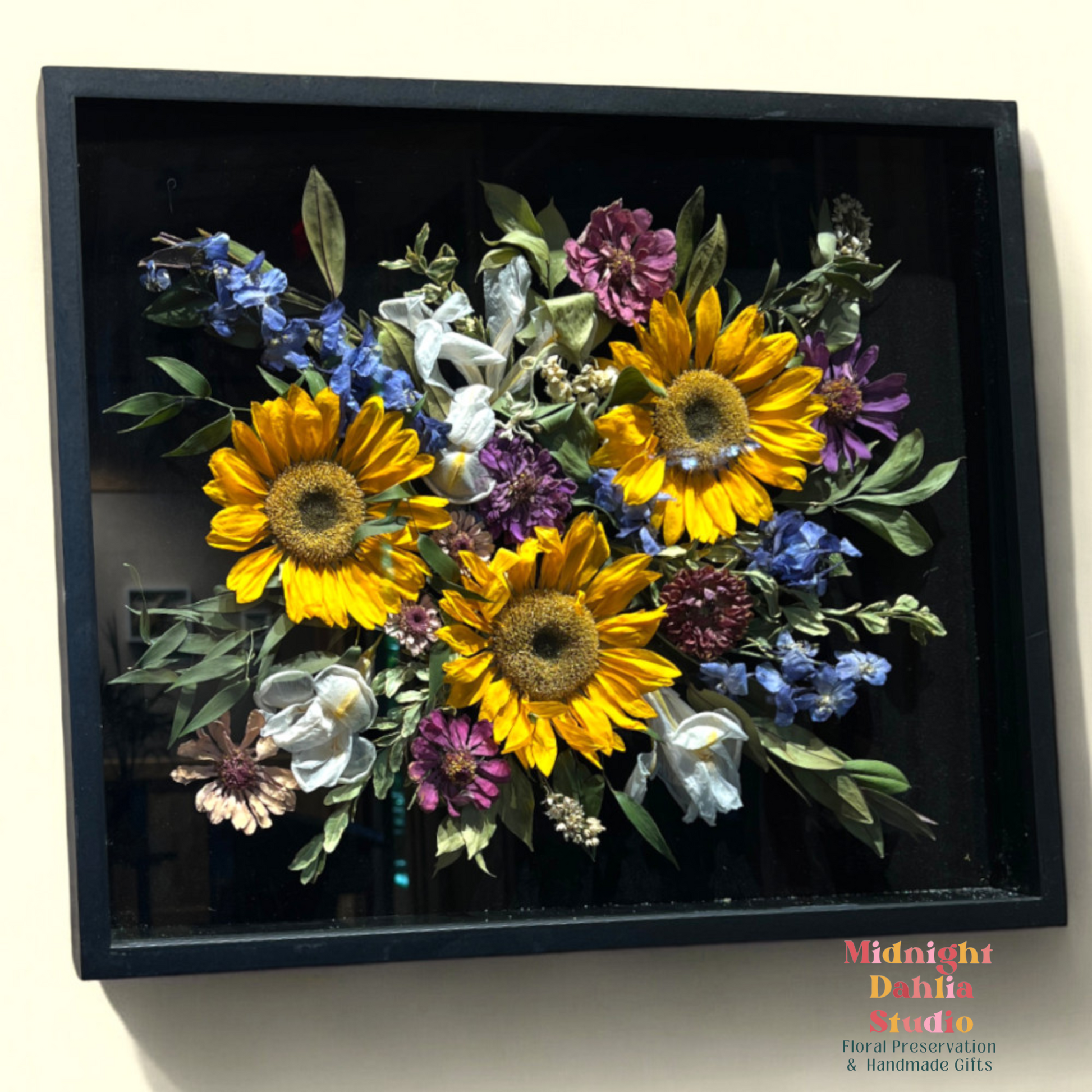 a wooden framed shadow box filled with many flowers, including yellow sunflowers, white lilies, purple zinnia, blue delphinium, and green leaves.