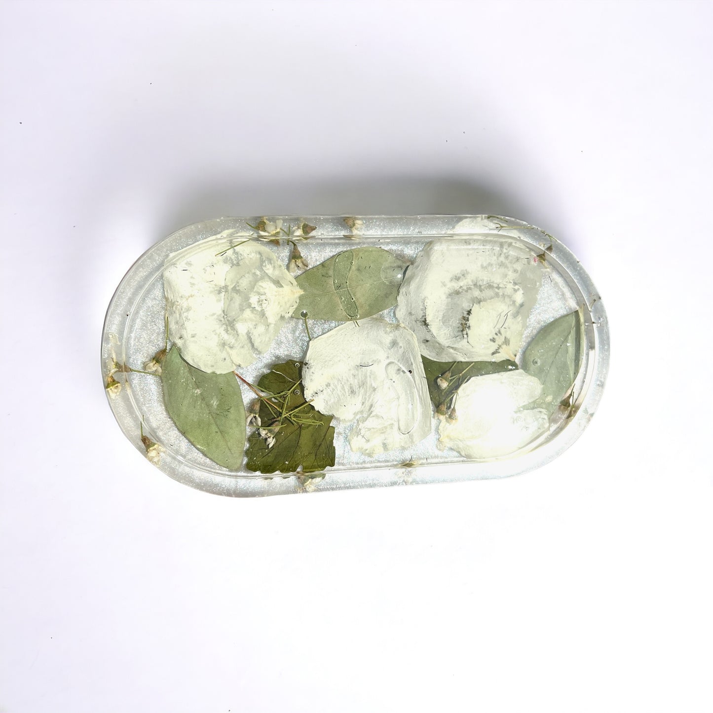 a clear resin vanity trinket tray with white rose petals and green leaves against a white background