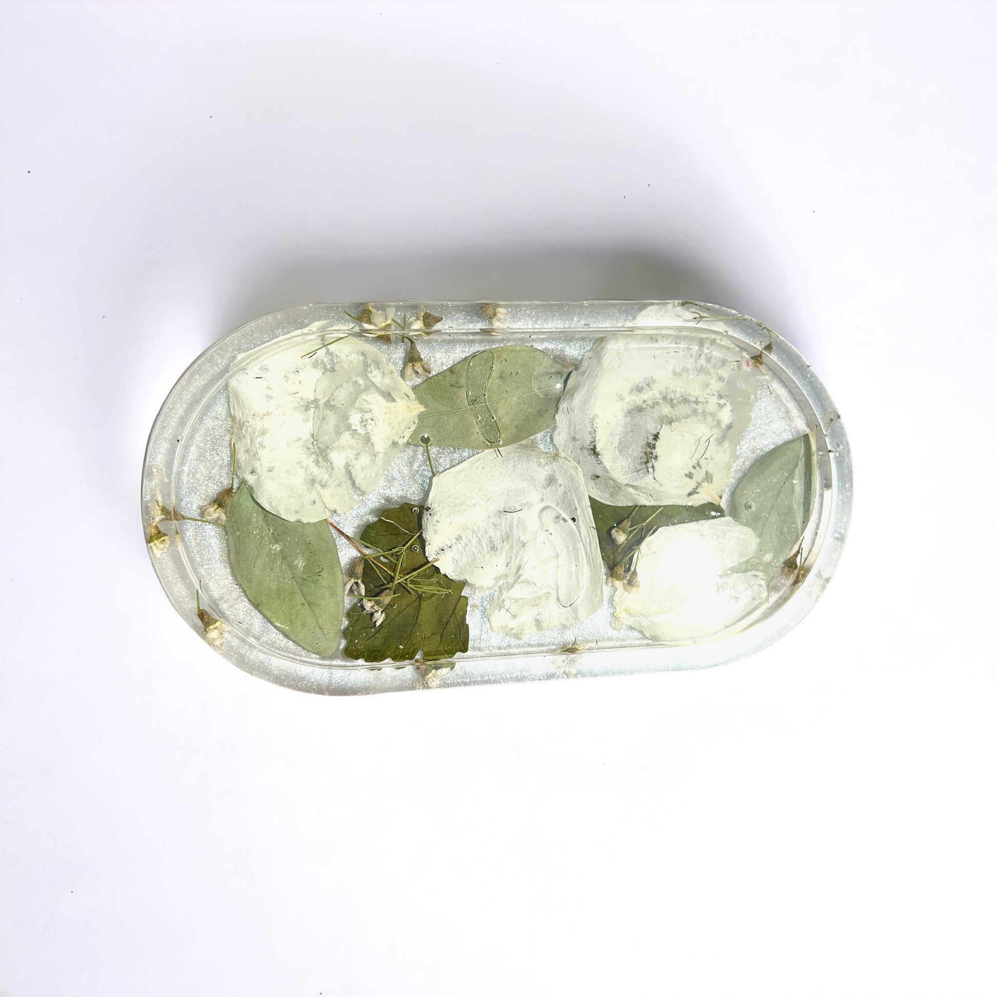a clear resin vanity trinket tray with white rose petals and green leaves against a white background