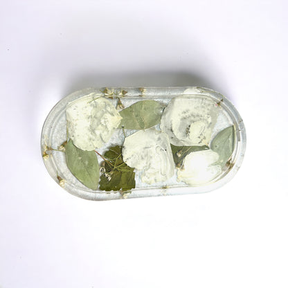 a clear resin vanity trinket tray with white rose petals and green leaves against a white background