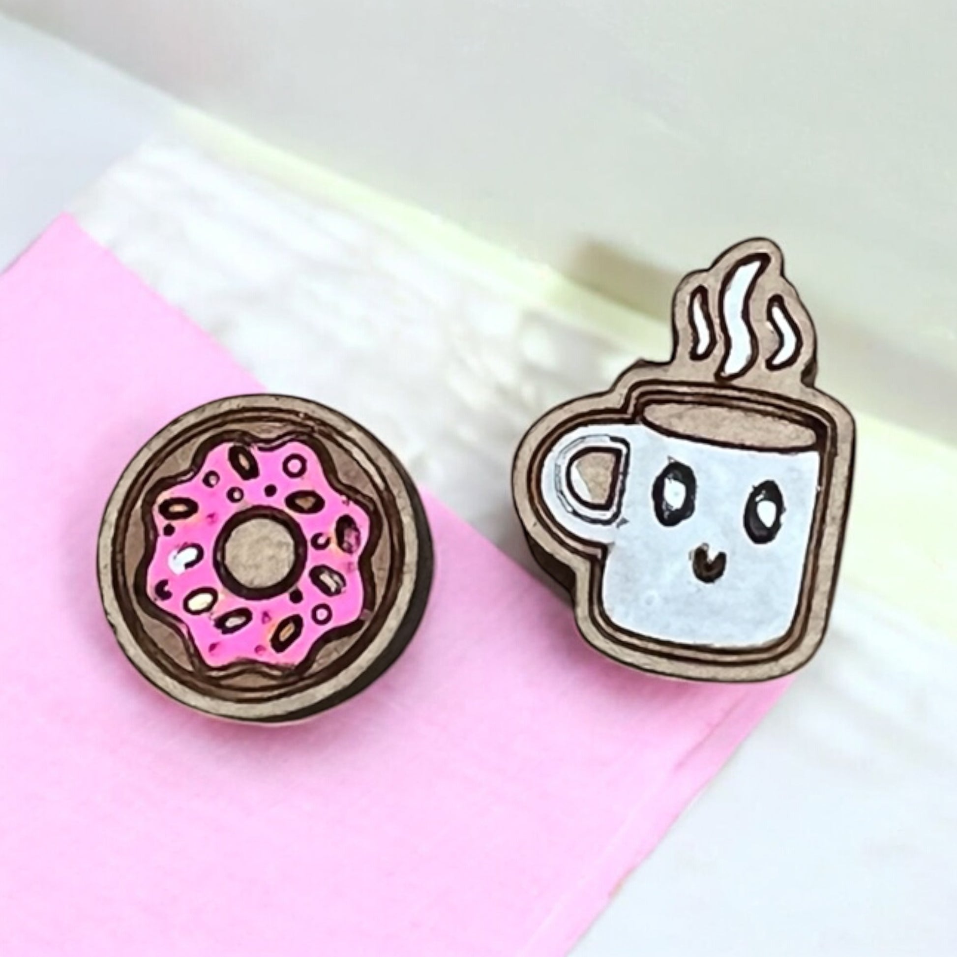 Two wooden earrings shaped like a donut and a coffee cup on a pink and white background.