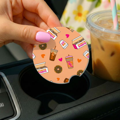 Circular drink coaster with donut and coffee design held over a cup in a car.