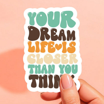 a white sticker that reads your dream life is closer than you think in bubble letters in teal blue green, brown, and mustard yellow