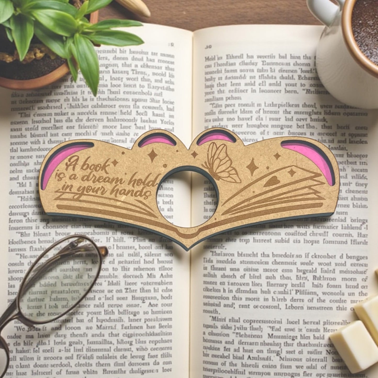 Wooden book-shaped object with pink interior on an open book, next to a cup of coffee and glasses.
