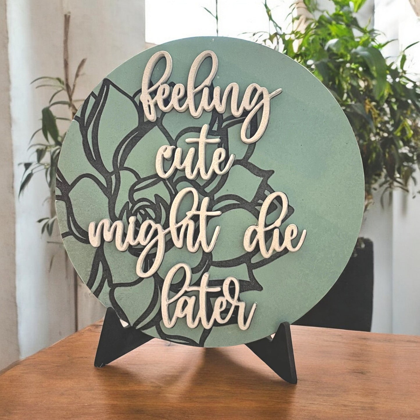 a round sign that is painted green with a black aloe plant stylistically engraved on the background with 3d words reading feeling cute might die later in a pretty cursive script.