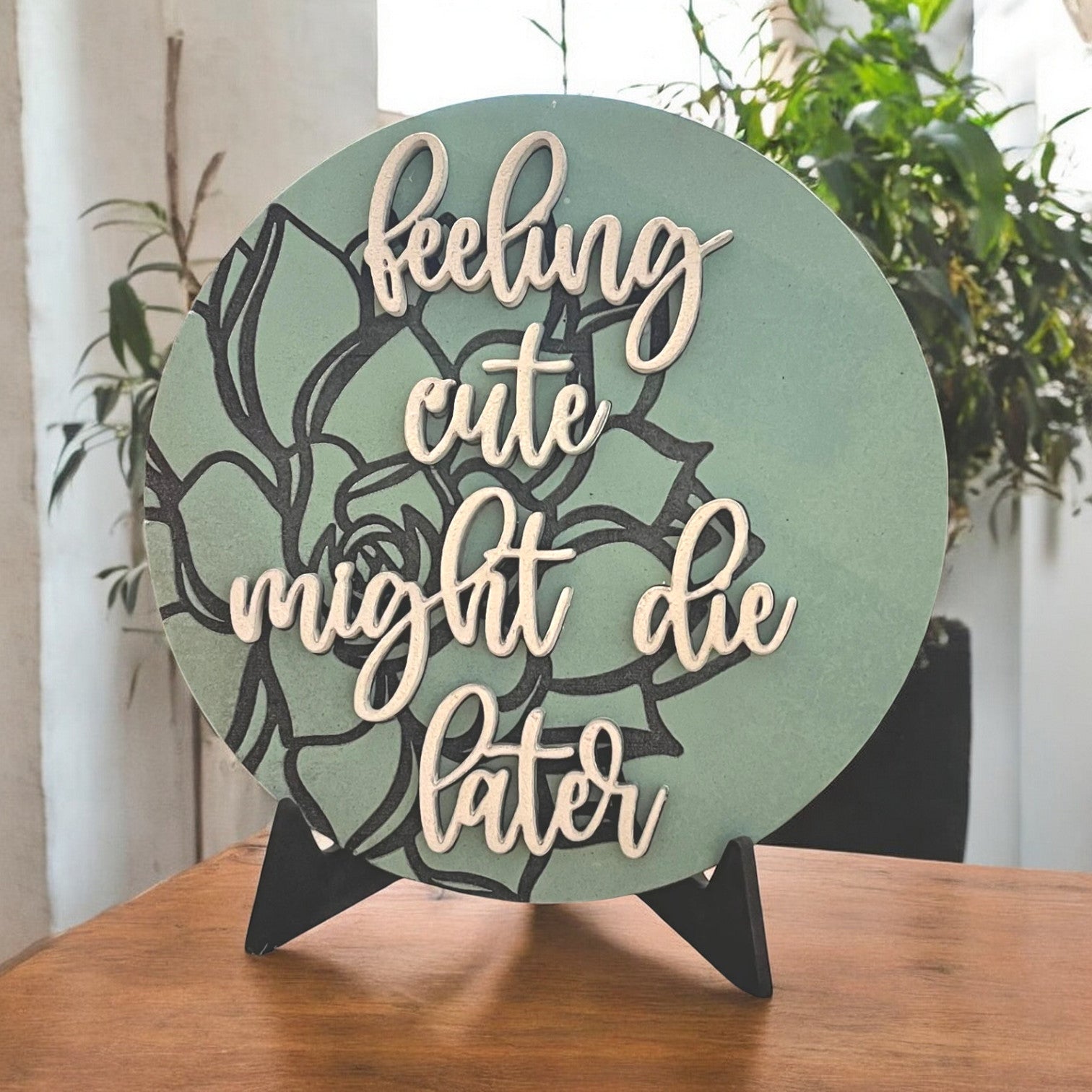 a round sign that is painted green with a black aloe plant stylistically engraved on the background with 3d words reading feeling cute might die later in a pretty cursive script.