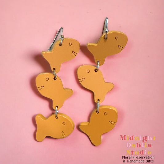 a pair of earrings with three orange goldfish cracker shapes with an engraved eye and smile, with silver-plated hardware.  a whimsical gift for women and moms.