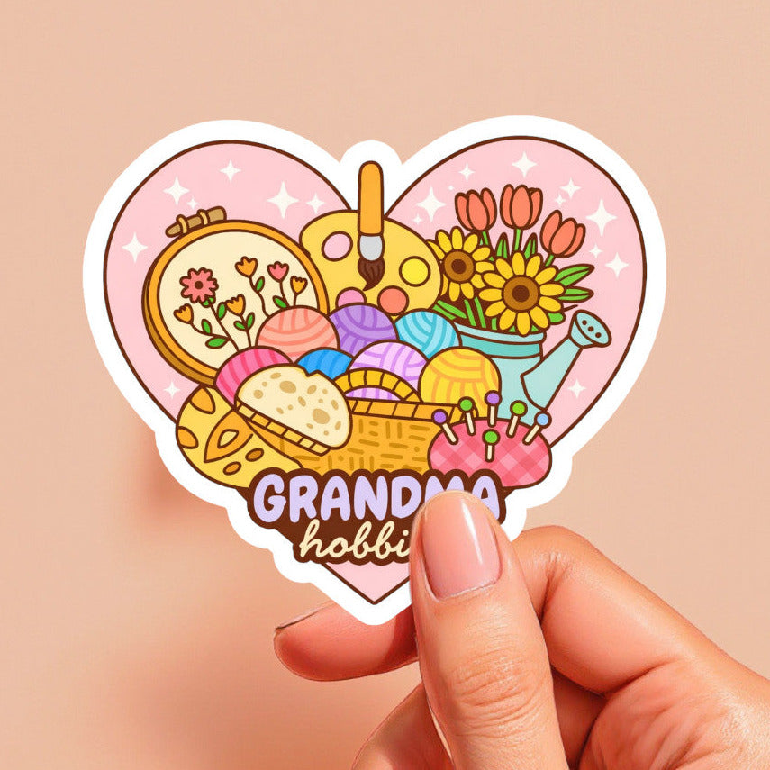Heart-shaped sticker with 'Grandma hobbies' design held by a hand on a beige background