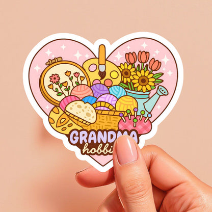 Heart-shaped sticker with 'Grandma hobbies' design held by a hand on a beige background