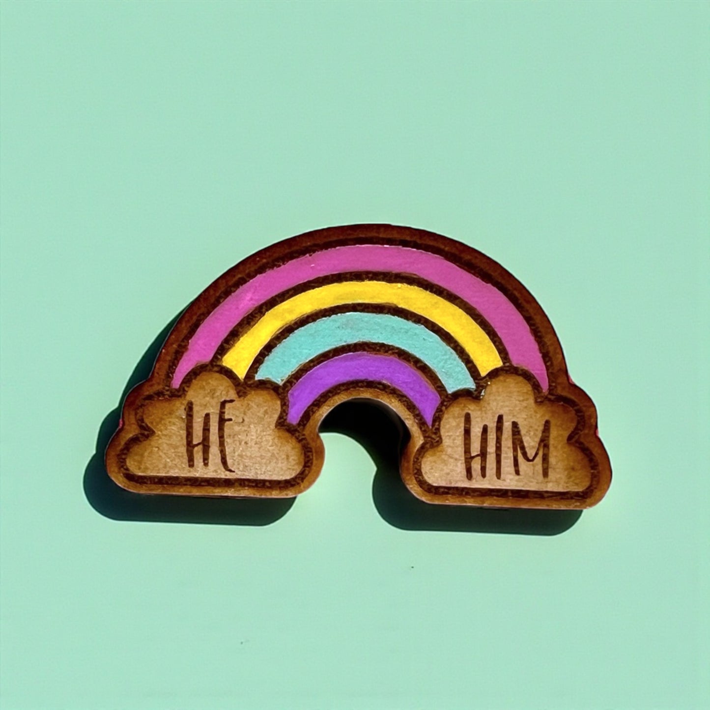 Wooden rainbow charm with 'HE' and 'HIM' on a light green background