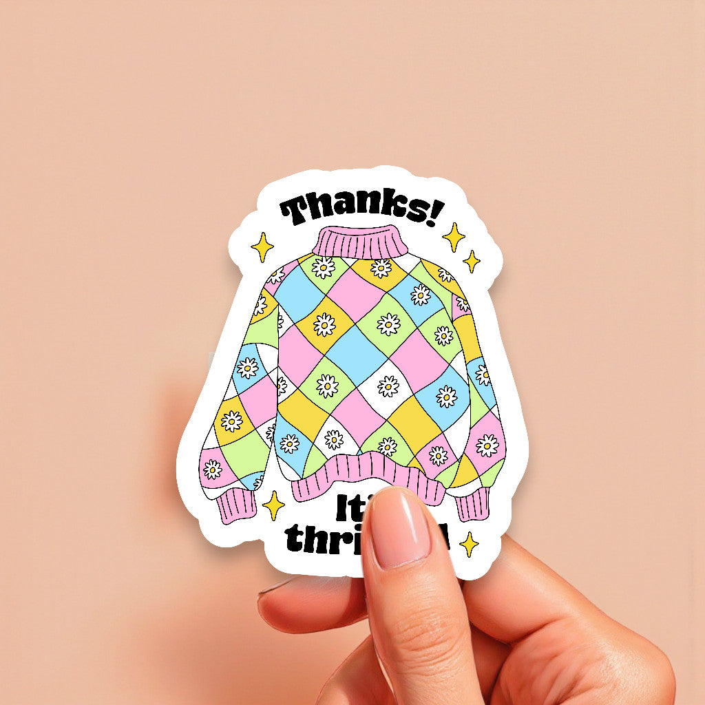 Colorful sweater-shaped sticker with 'Thanks! It's thrifted!' text held by a hand on a beige background
