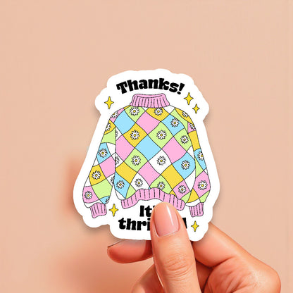 Colorful sweater-shaped sticker with 'Thanks! It's thrifted!' text held by a hand on a beige background