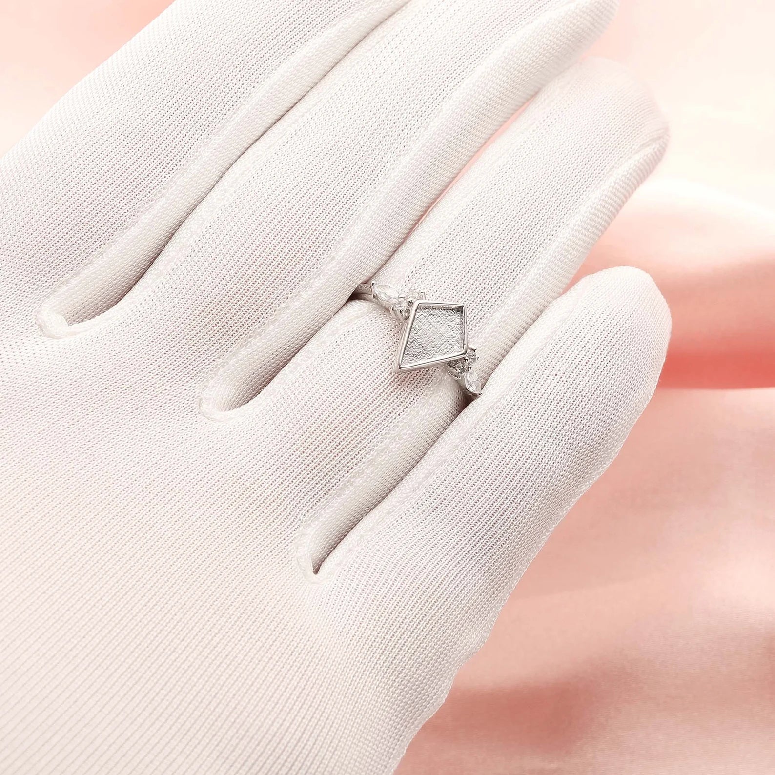 Silver ring with a diamond-shaped stone on a white glove against a soft pink background