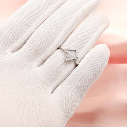 Silver ring with a diamond-shaped stone on a white glove against a soft pink background