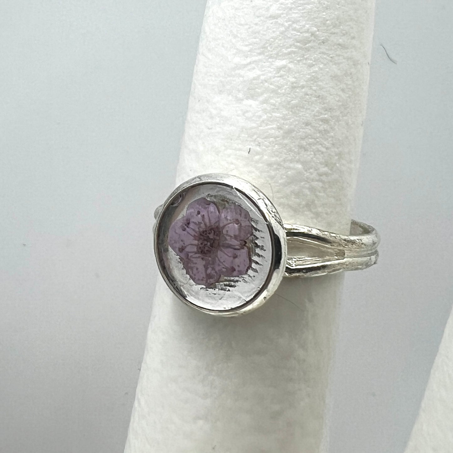 Silver ring with a purple forget me not real pressed flower on a white cylindrical object against a gray background