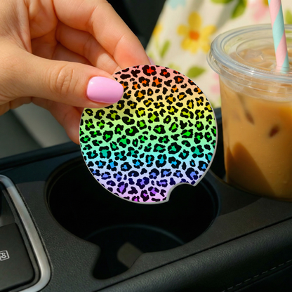 Hand holding a rainbow leopard print car coaster over a cup holder with a drink in the background.
