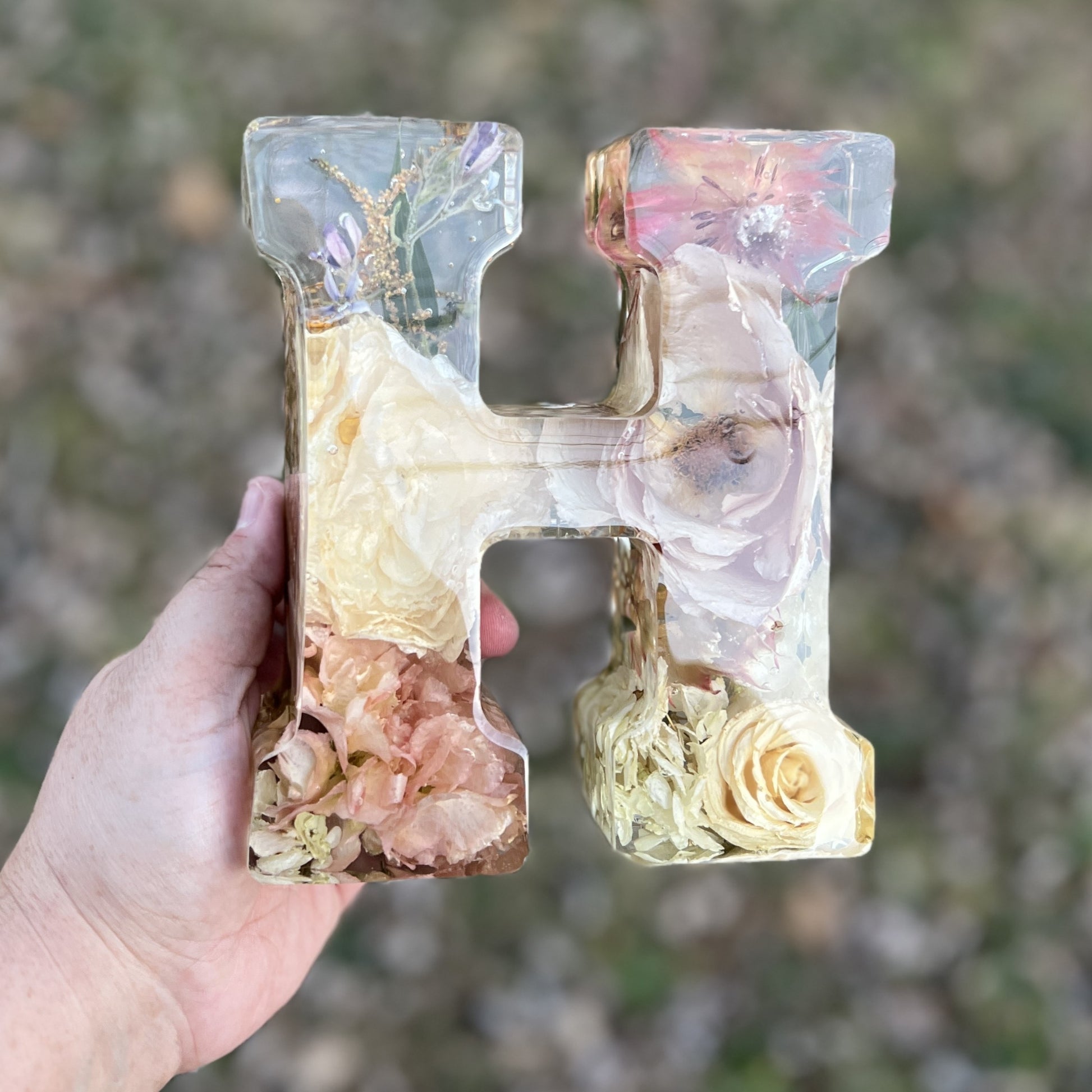 Decorative letter 'H' with floral elements held by a hand against a blurred natural background