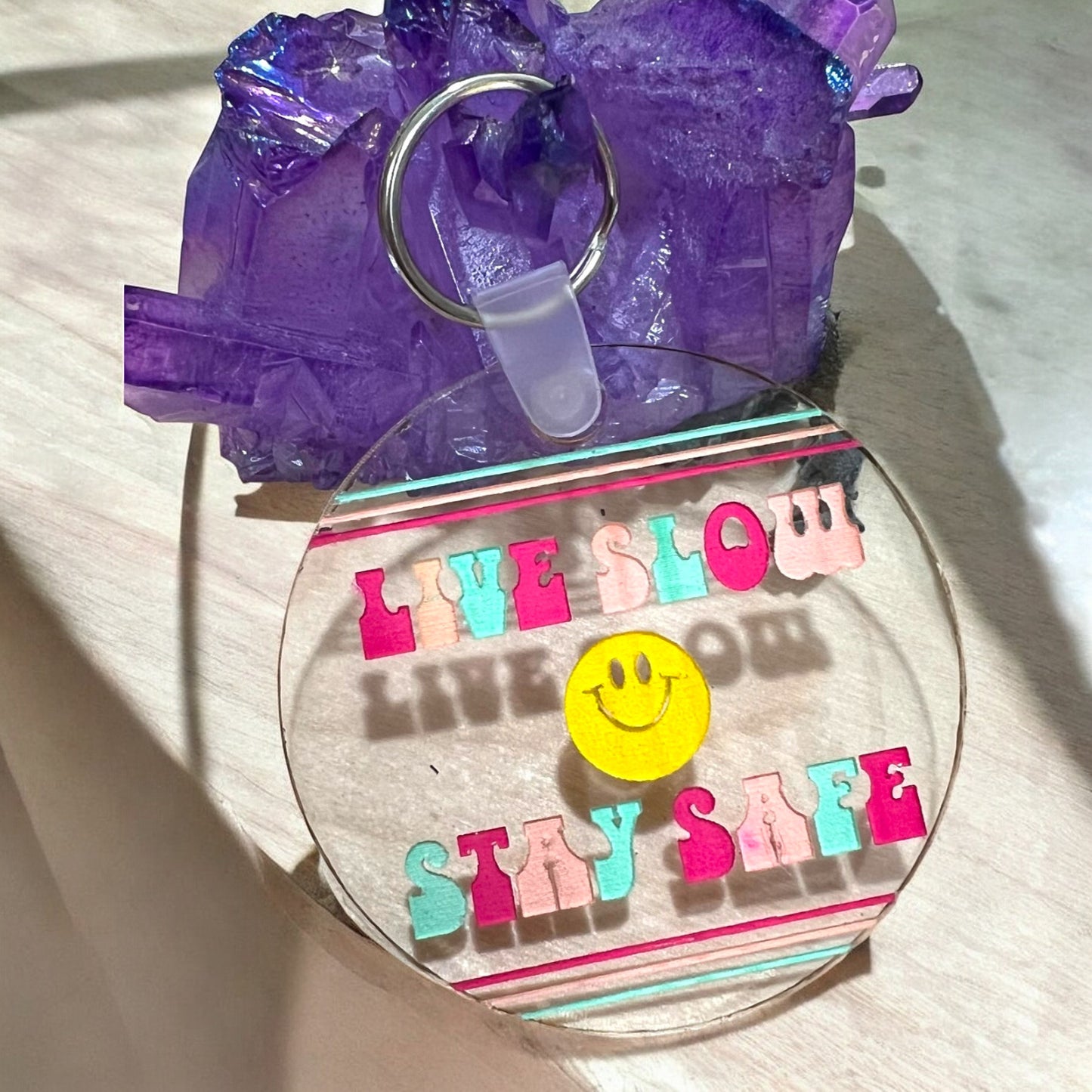 a round clear acrylic keychain that reads live slow stay safe with a smiley smile face in the middle and lines on the top and bottom of the keychain, colors are pink, peach orange, and teal blue green.