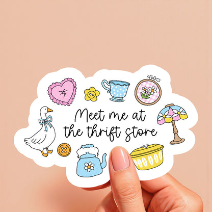 Sticker with illustrations and text 'Meet me at the thrift store' held by a hand on a beige background