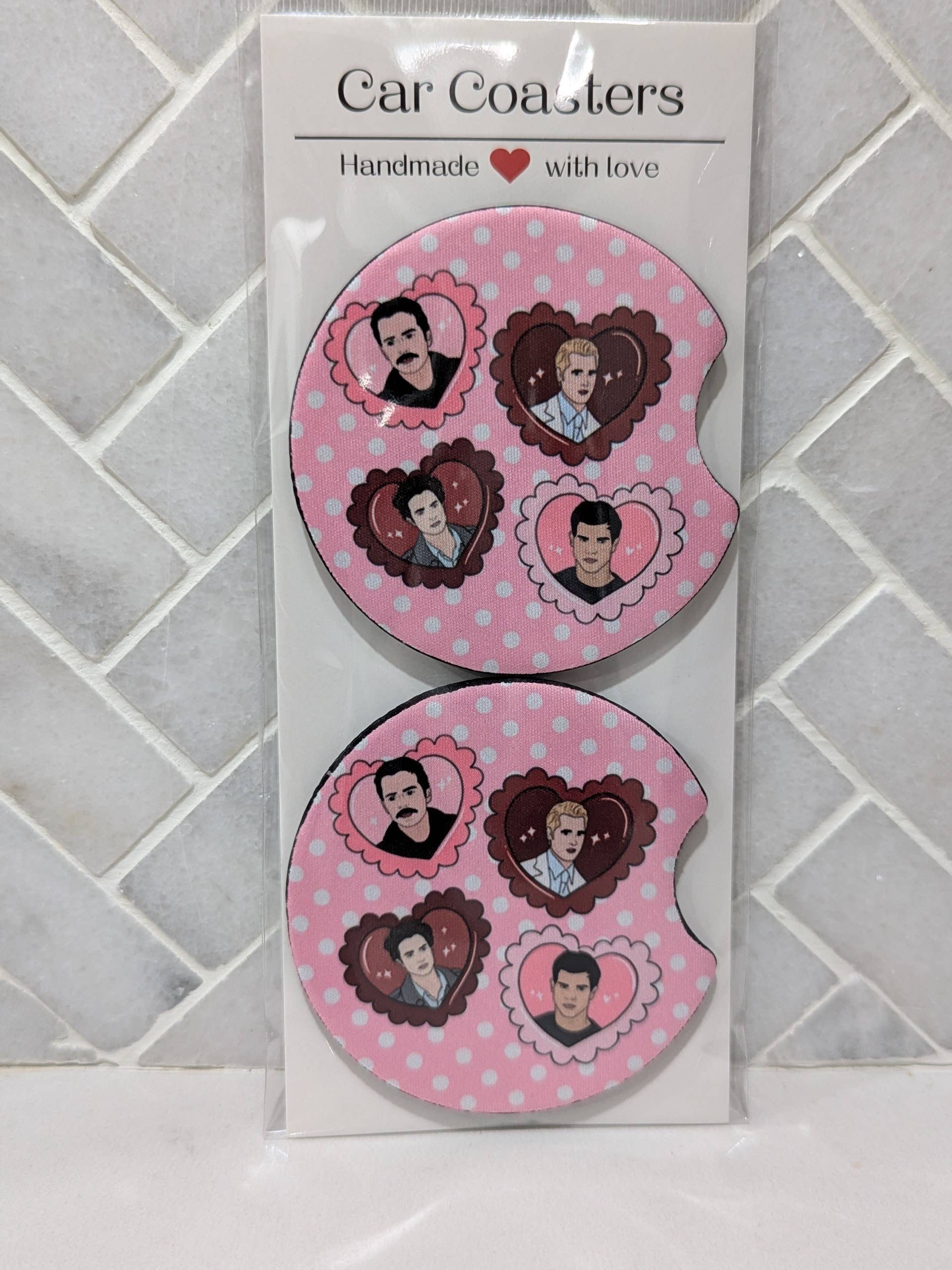 Two pink car coasters with heart designs on a white card against a tiled wall background.