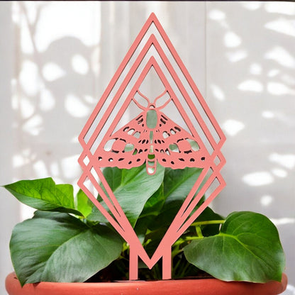 a plant trellis made of peach acrylic in a diamond shape with a moth in the center, in a plant