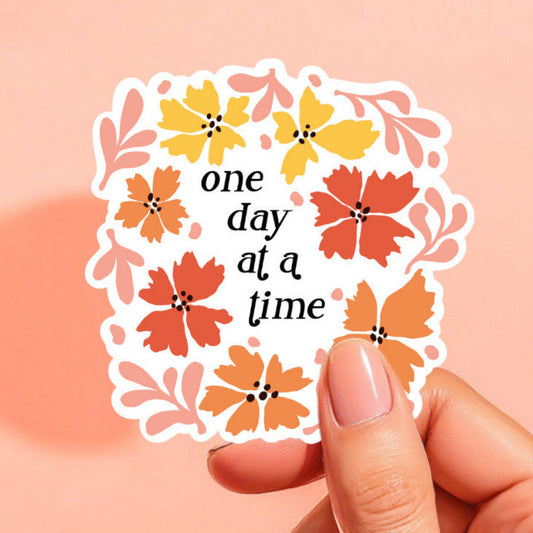 a white sticker that reads one day at a time in black surrounded by flowers