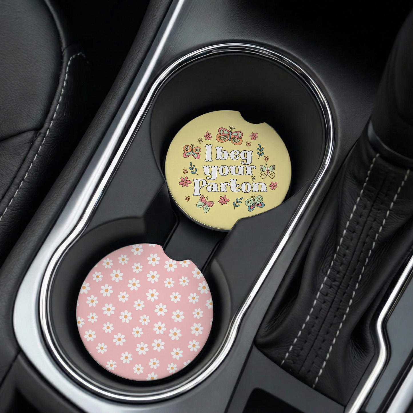 a set of car coasters in car cup holders, one pink with floral pattern and the other yellow with a humorous phrase