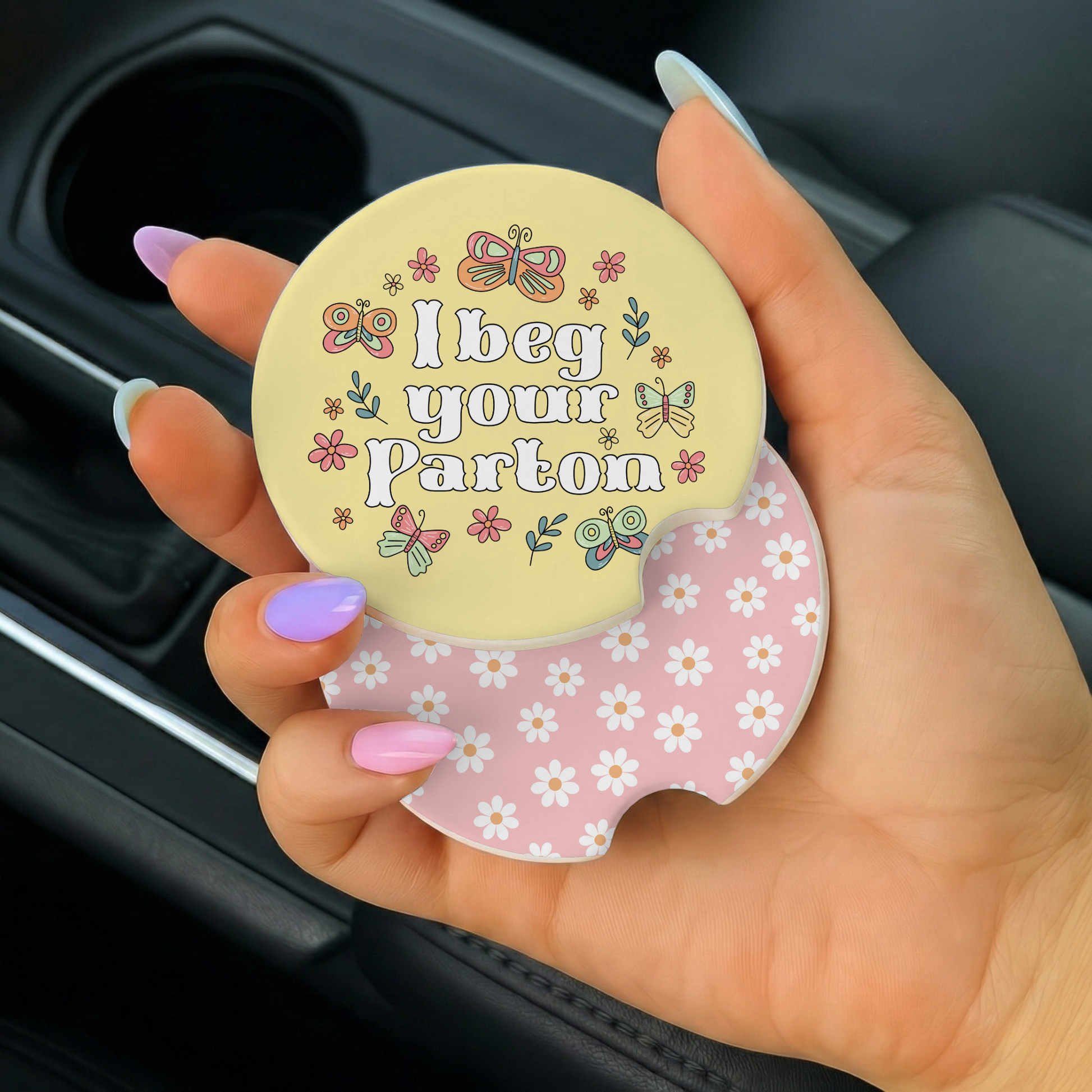 Hand holding a car coaster with 'I beg your pardon' text and floral design.