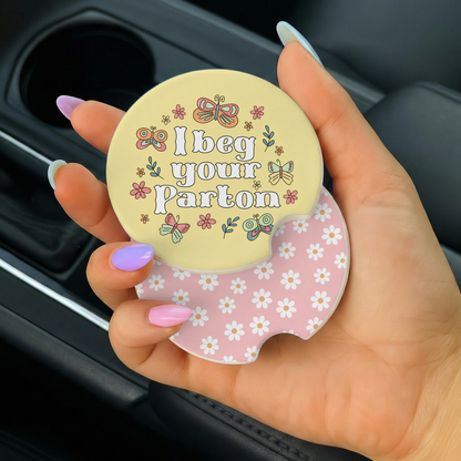 Hand holding a car coaster with 'I beg your pardon' text and floral design.