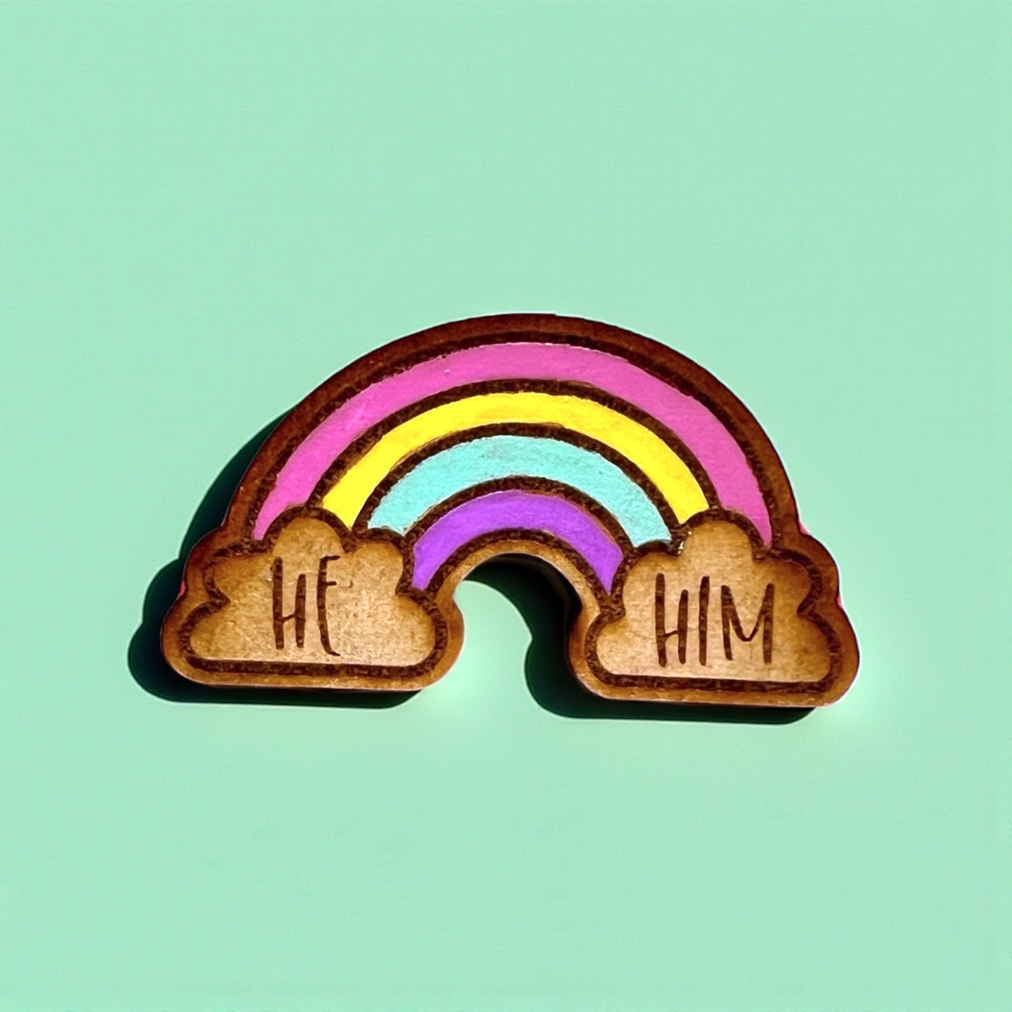 Wooden rainbow charm with 'HE' and 'HIM' on clouds against a teal background