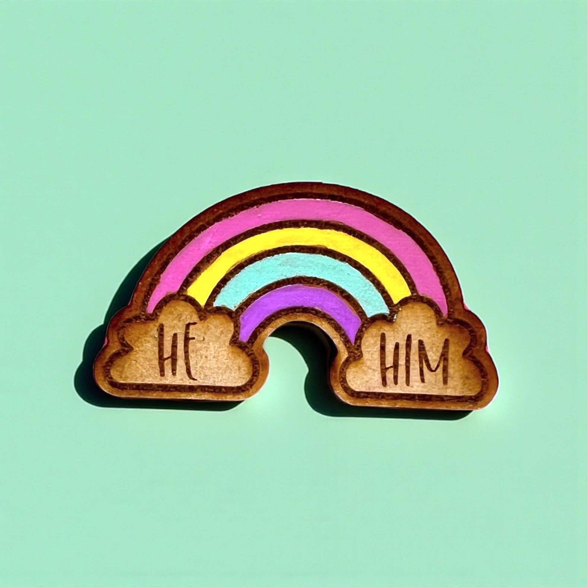 Wooden rainbow charm with 'HE' and 'HIM' on clouds against a teal background