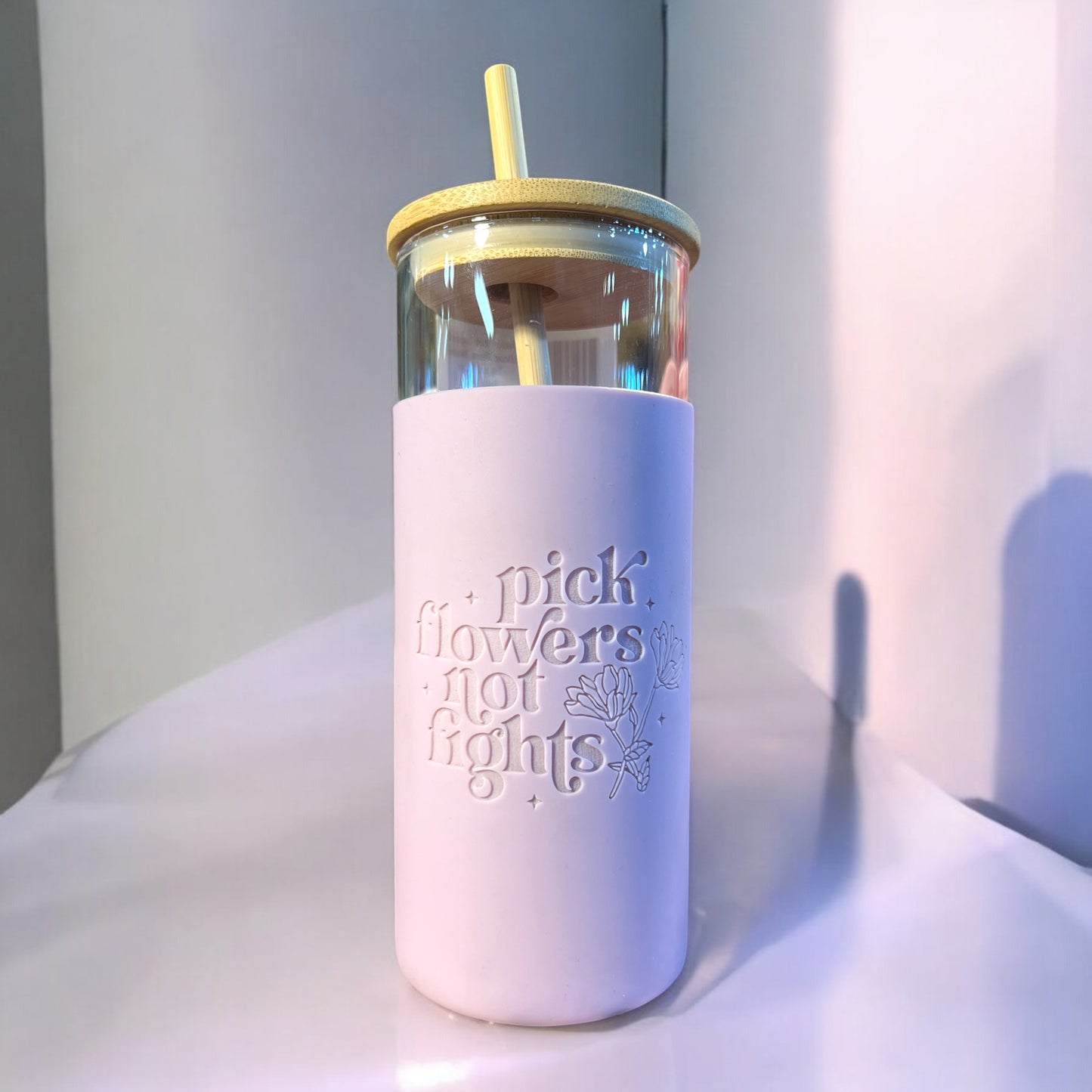 A pink tumbler with a clear glass body, a silicone engraved floral design, a bamboo straw, and the text 'PICK FLOWERS NOT FIGHTS'.