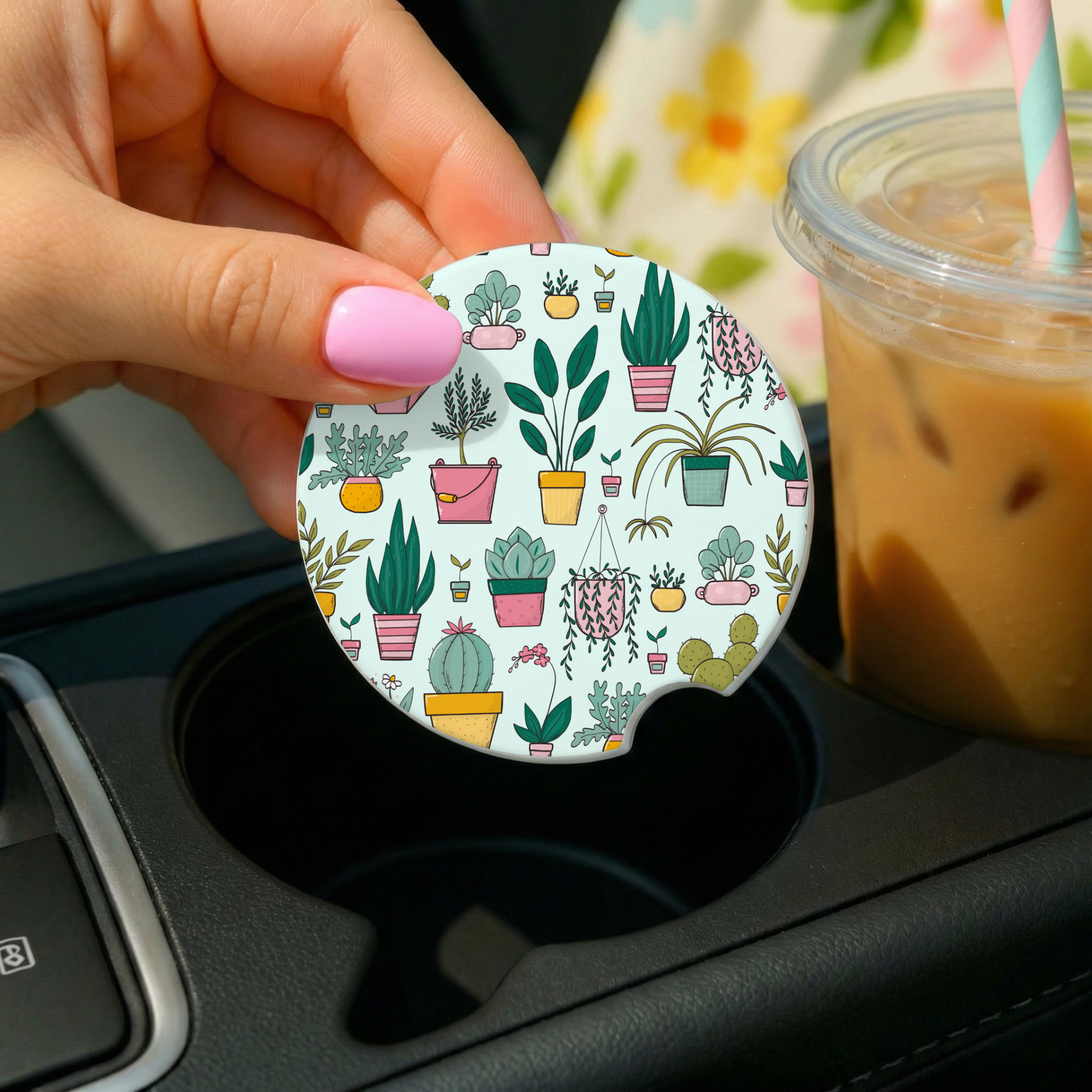 Hand holding a car coaster with plant pattern next to a drink in a car cup holder.