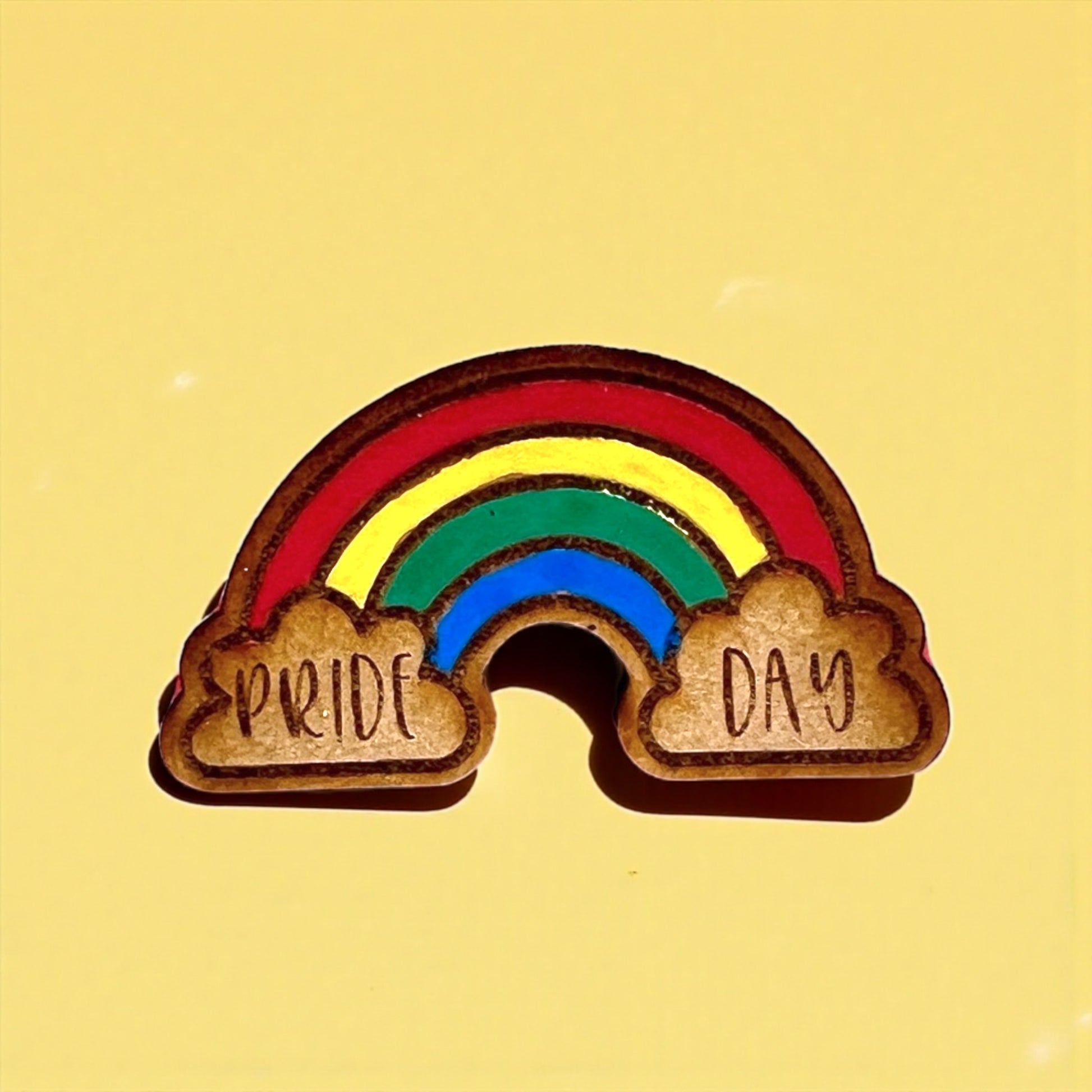 Wooden rainbow pin with 'Pride Day' on a yellow background