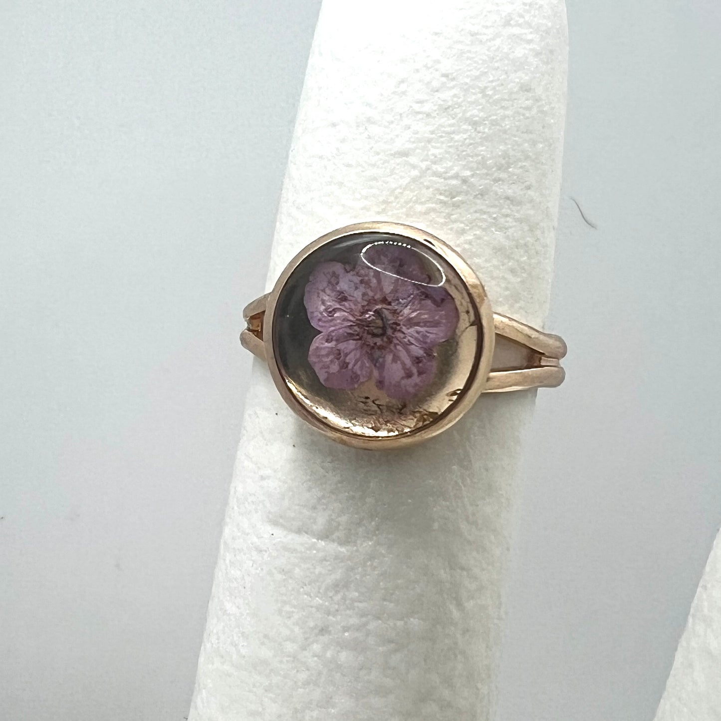 Gold ring with a purple forget me not real pressed flower on a white cushion against a light gray background