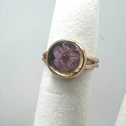 Gold ring with a purple forget me not real pressed flower on a white cushion against a light gray background