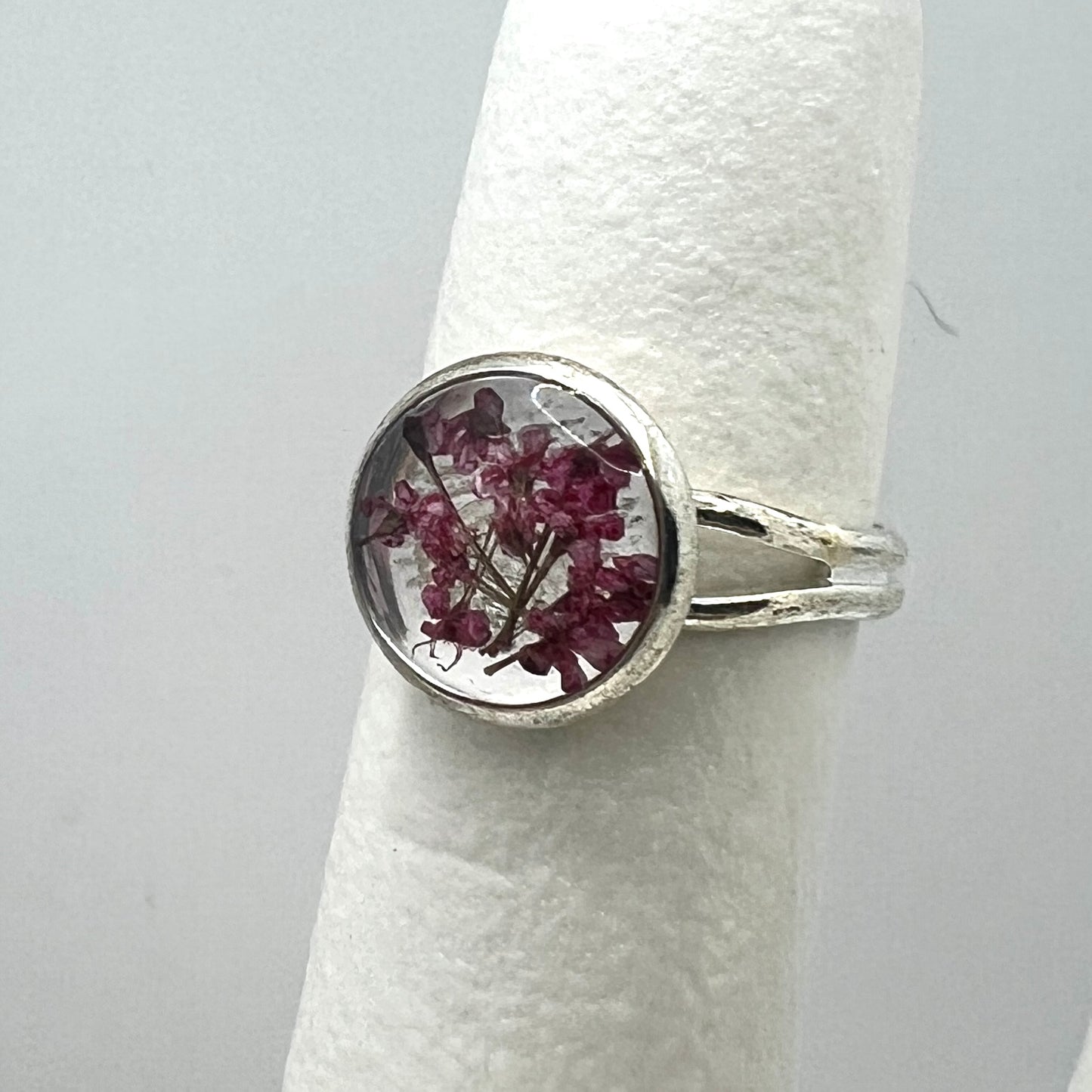 Silver ring with a circular design featuring dried flowers on a white cylindrical stand against a light gray background.