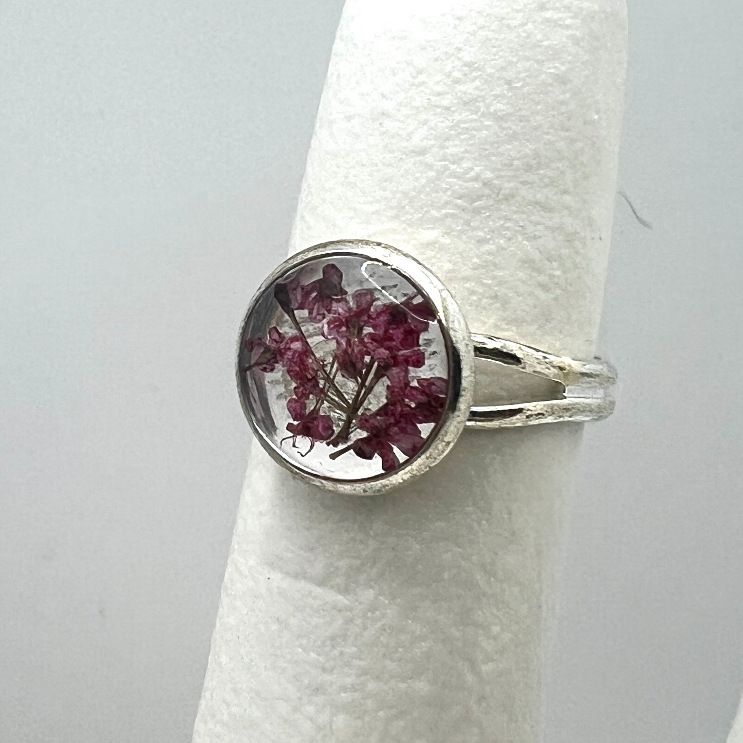 Silver ring with a circular design featuring dried flowers on a white cylindrical stand against a light gray background.