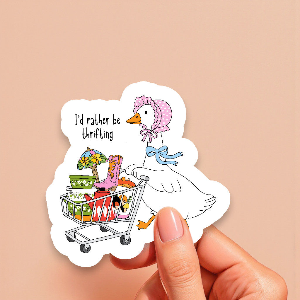 Sticker of a duck with a shopping cart full of items and 'I'd rather be thrifting' text, held by a hand against a beige background.
