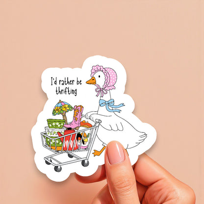 Sticker of a duck with a shopping cart full of items and 'I'd rather be thrifting' text, held by a hand against a beige background.