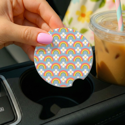 Hand holding a circular rainbow-patterned car coaster over a cup holder with a drink in the background.